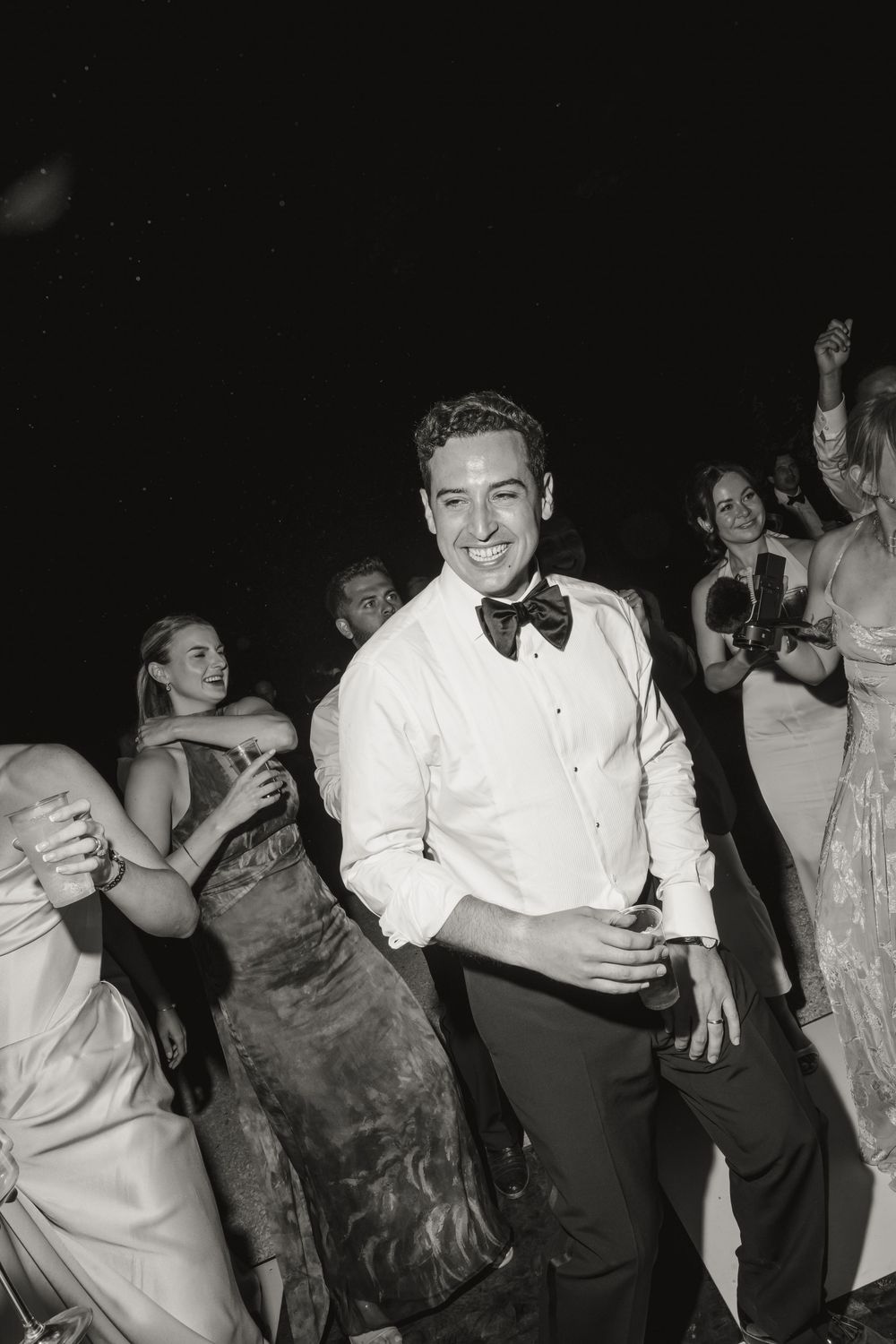 Smiling groom in white tuxedo shirt and bow tie dancing with guests at wedding reception.