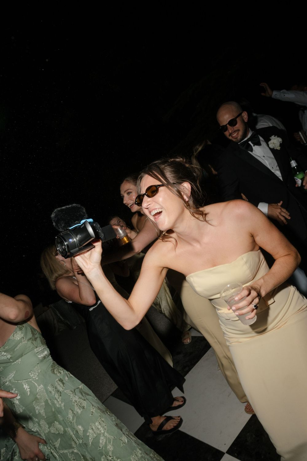 Bridesmaid in yellow strapless dress films wedding reception dancing on checkered floor wearing sunglasses.