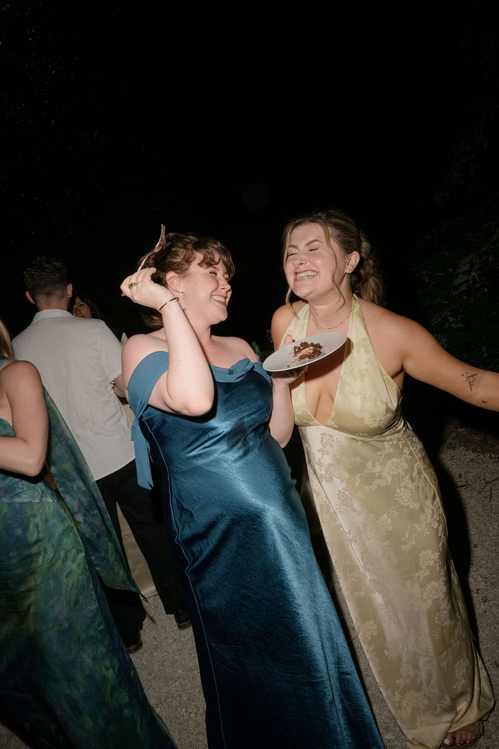 Two women in elegant satin gowns laughing and dancing at a formal outdoor wedding reception.