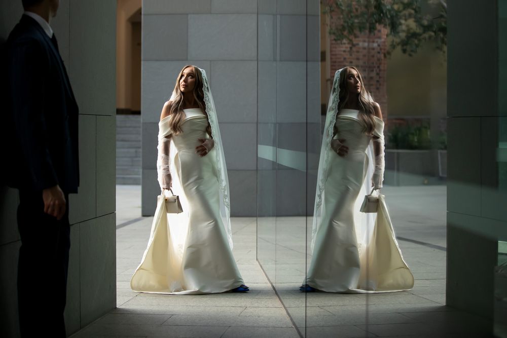 Bride in white gown and veil reflected in mirror, standing beside groom in dark suit in elegant venue.