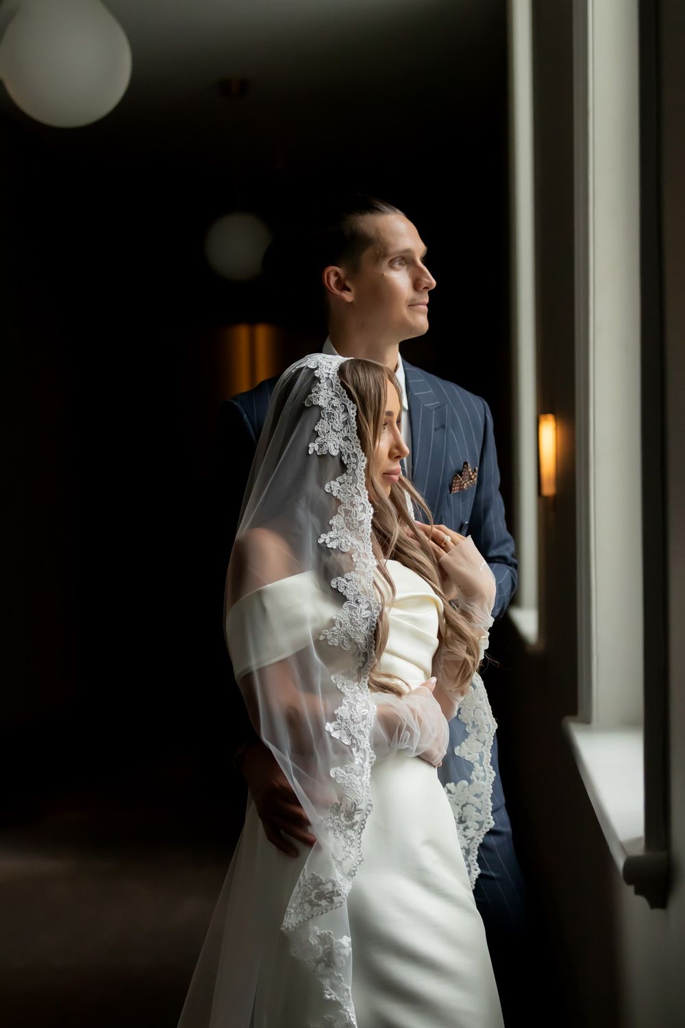 Bride in lace veil and groom in blue suit standing by window in elegant indoor wedding portrait.