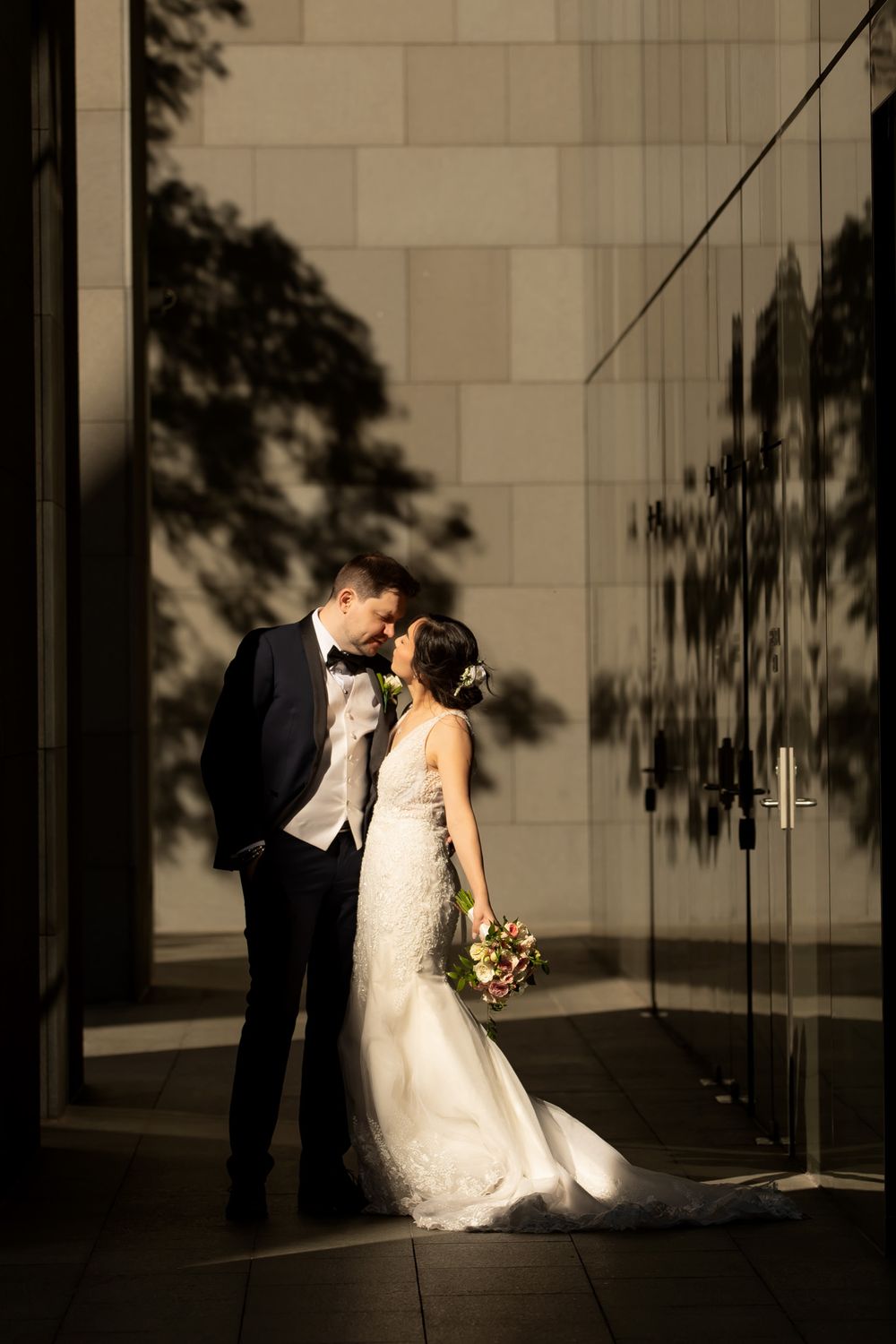 Bride and groom share a romantic moment in golden light against a modern stone wall, bride holding a colorful bouquet.