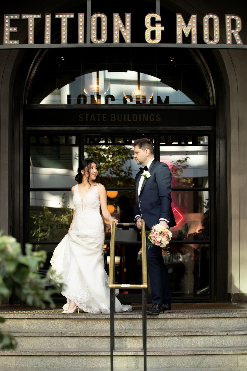 Bride and groom stand on steps outside Long Chim State Buildings, bride in white gown, groom in navy tuxedo holding bouquet.