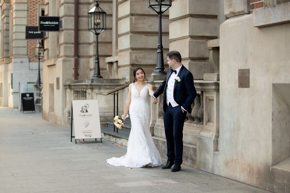 Bride and groom holding hands walking along a classic stone building on their wedding day.