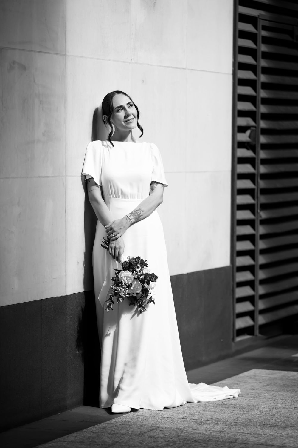 Black and white photo of a bride in a white gown holding a bouquet, leaning against a wall.
