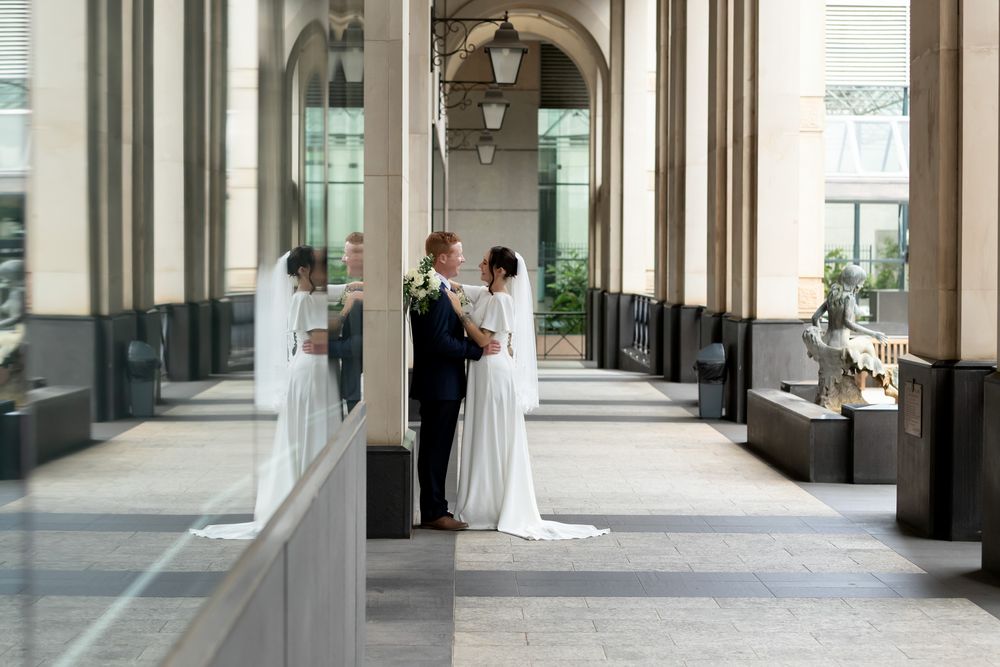 Bride and groom share a romantic moment in an elegant columned hallway, with their reflection visible in nearby glass.