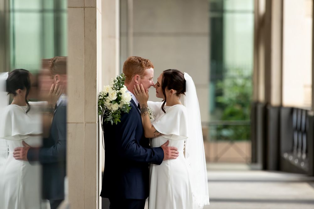 A bride and groom share a romantic moment beside a pillar, with the bride holding a white bouquet on their wedding day.