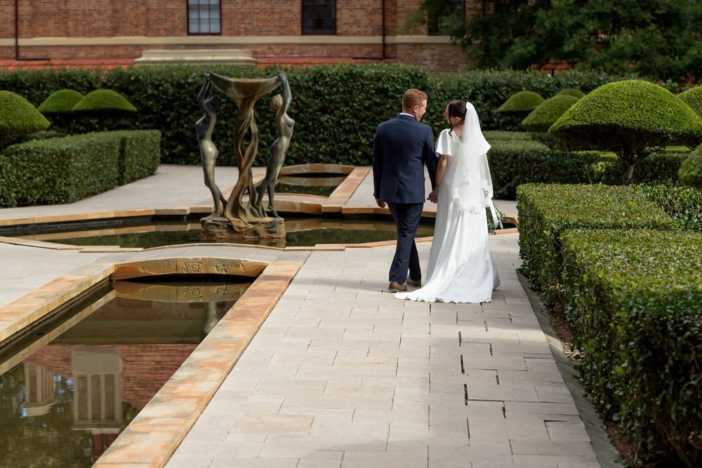 Bride and groom walking hand in hand through elegant formal garden with topiary hedges and ornamental fountain.