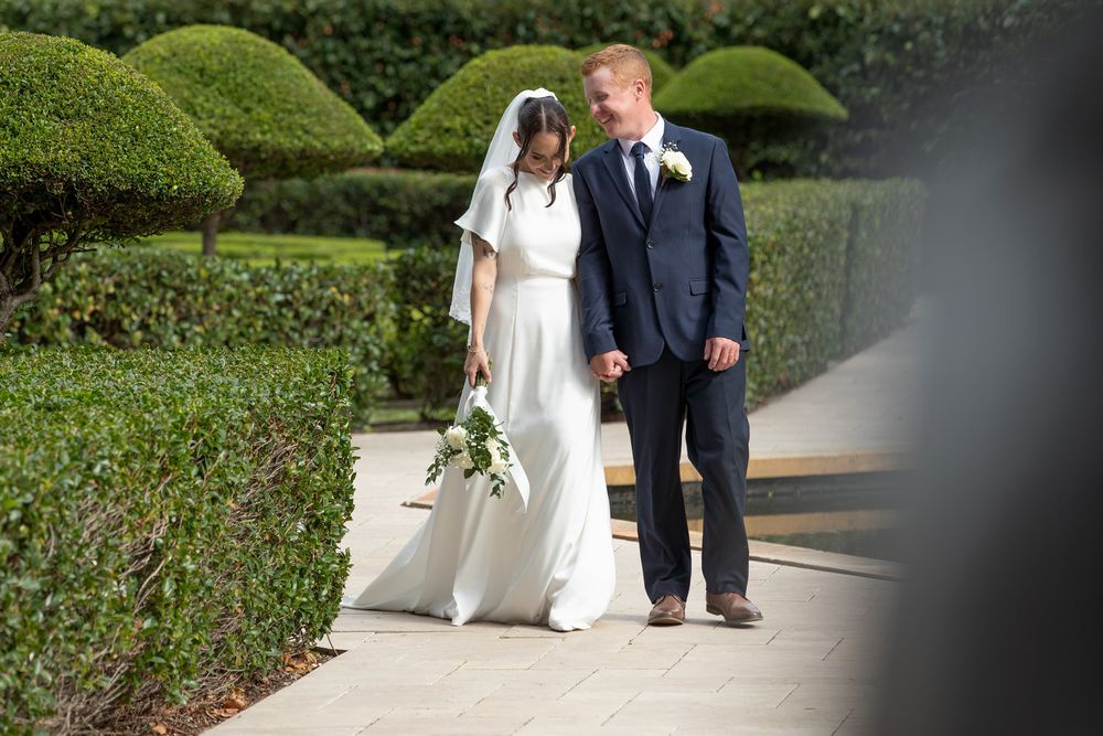 Bride and groom walking hand in hand through a formal garden with manicured hedges on their wedding day.