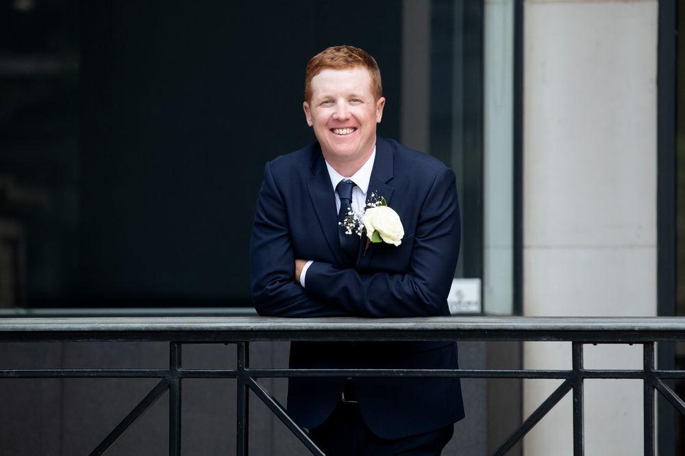 Smiling redhead groom in navy suit with boutonniere leaning on balcony railing at wedding venue.