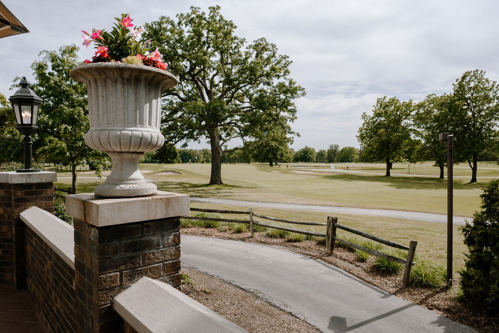 Stone urn planter with pink flowers on brick pillar overlooking lush green golf course with trees and pathway.
