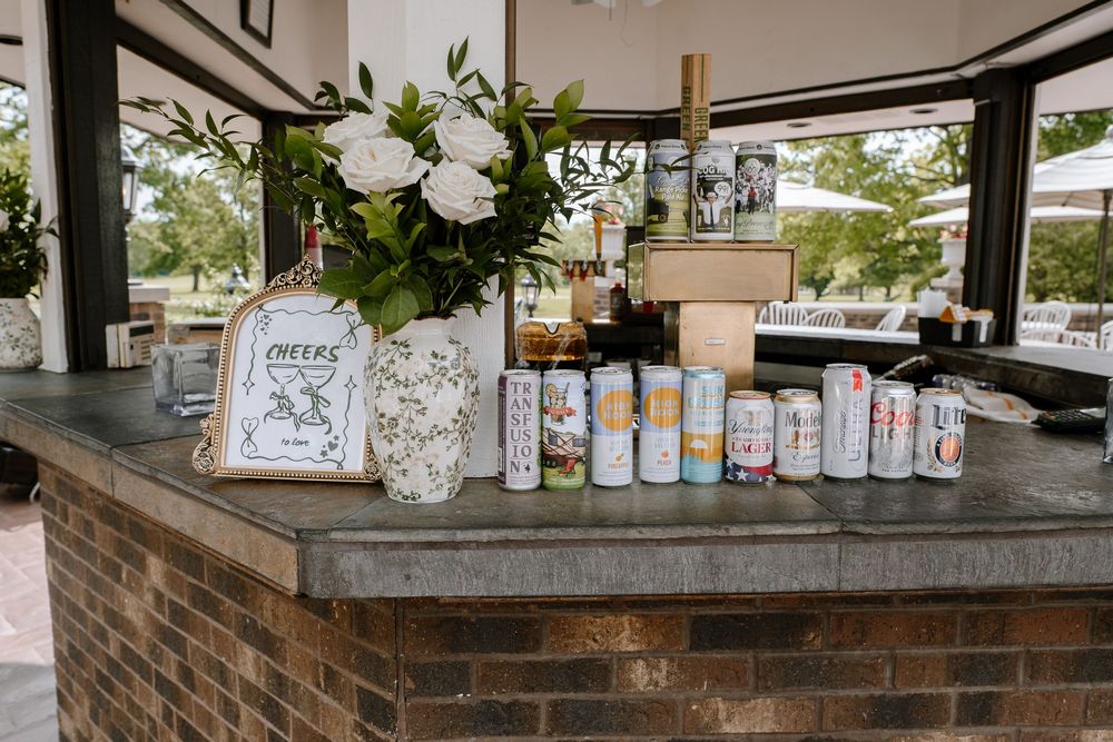 Outdoor bar display with canned beverages, white floral arrangement, and a Cheers sign at a wedding reception.