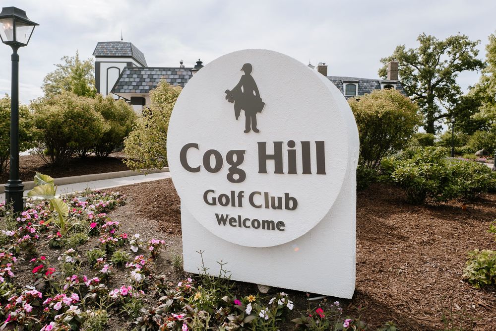 Cog Hill Golf Club welcome sign surrounded by colorful flowers and lush greenery.