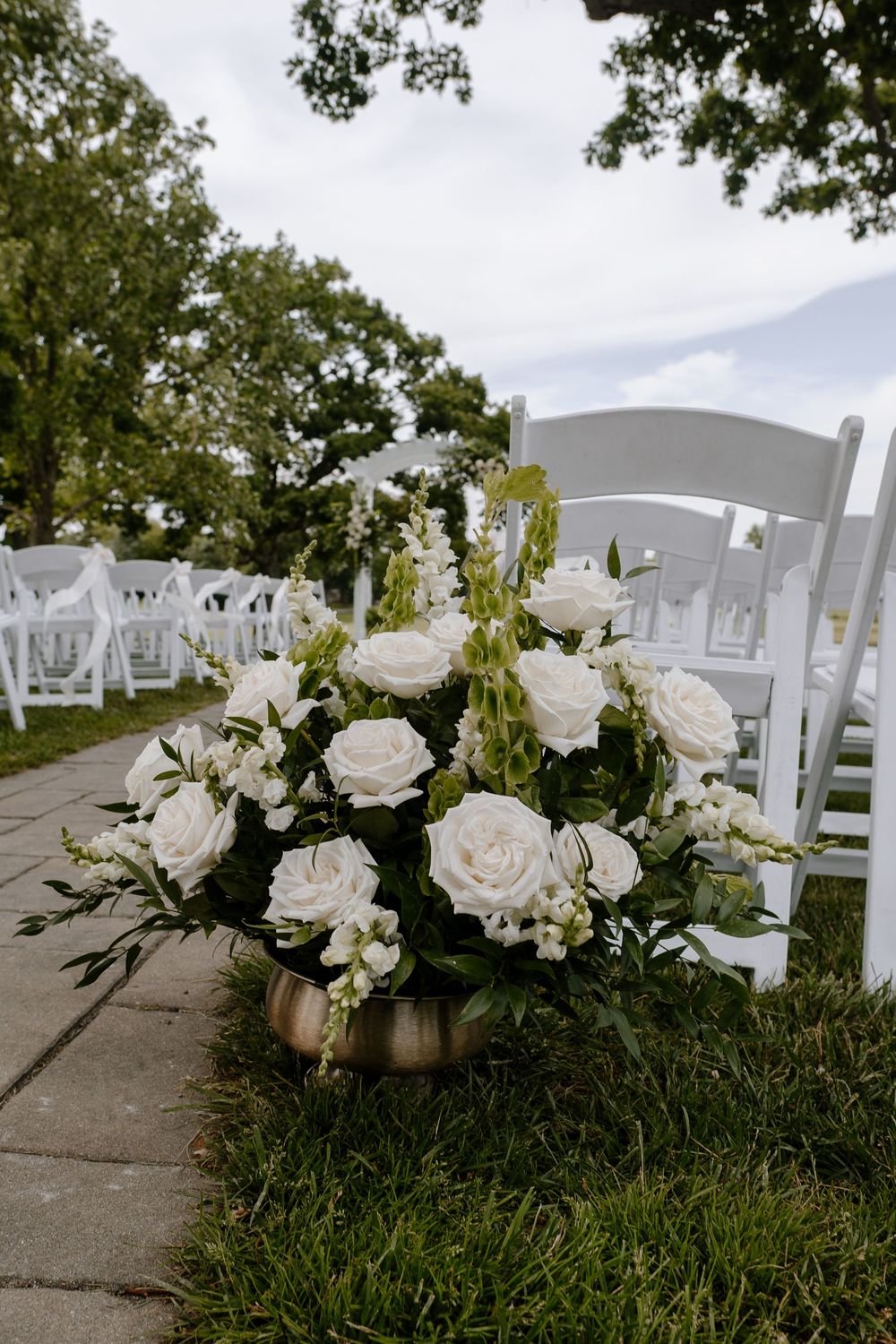 White roses and greenery in a gold vase line the aisle at an outdoor wedding ceremony with white chairs.