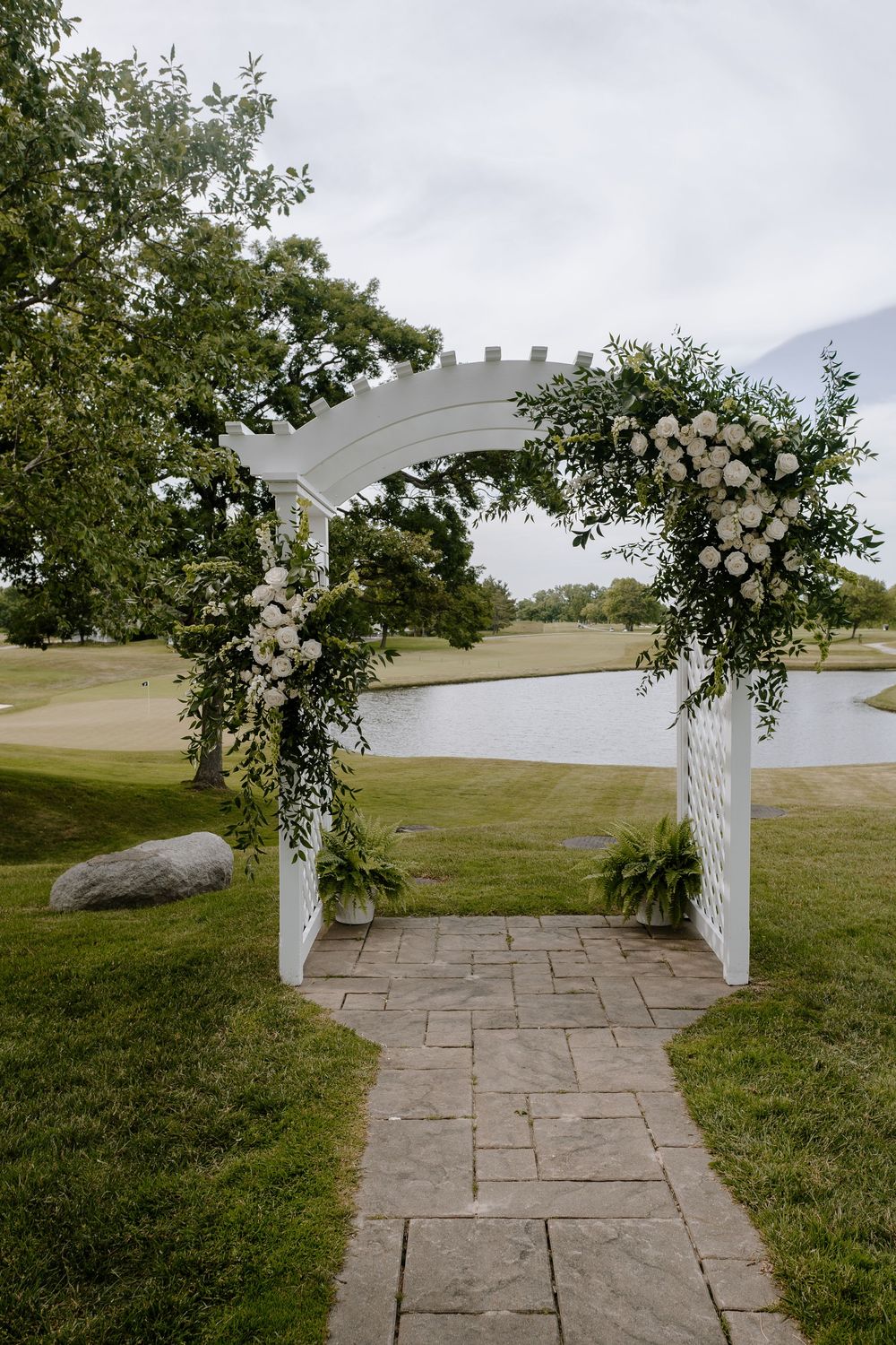 White wedding arch adorned with white roses and greenery on a stone path overlooking a serene pond.