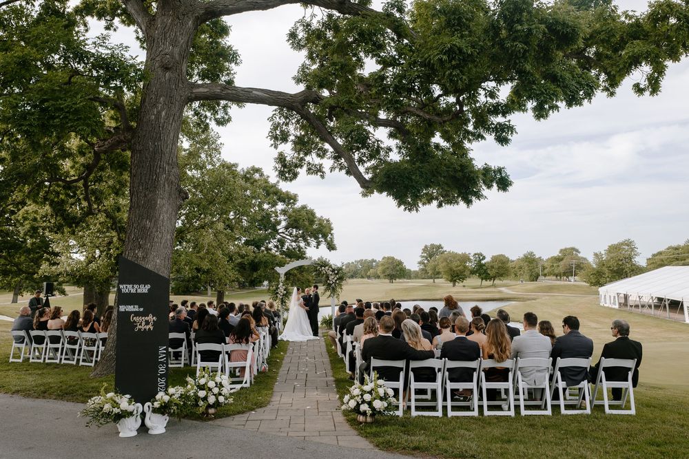 Outdoor wedding ceremony under a large oak tree with white chairs and floral arrangements on a lush green lawn.