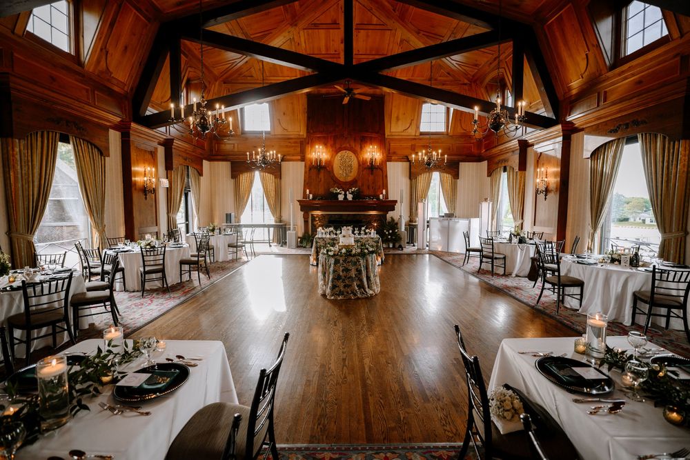 Elegant wedding reception hall with wood-beamed ceilings, chandelier lighting, round tables, and a grand fireplace centerpiece.