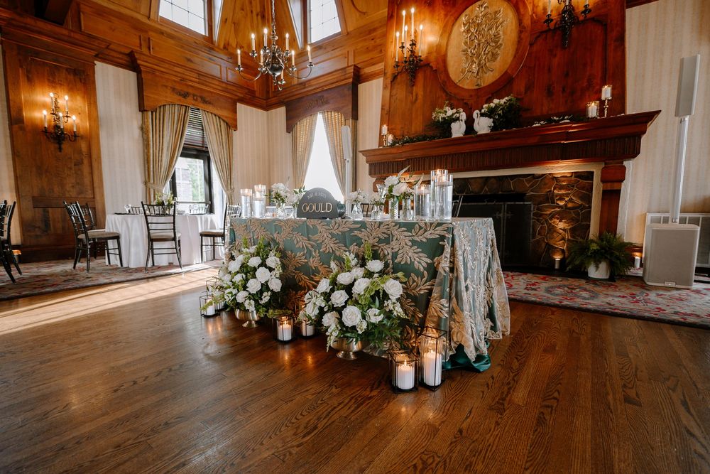 Elegant wedding sweetheart table with floral tablecloth, white roses, and candles in a rustic wood-paneled venue with fireplace.