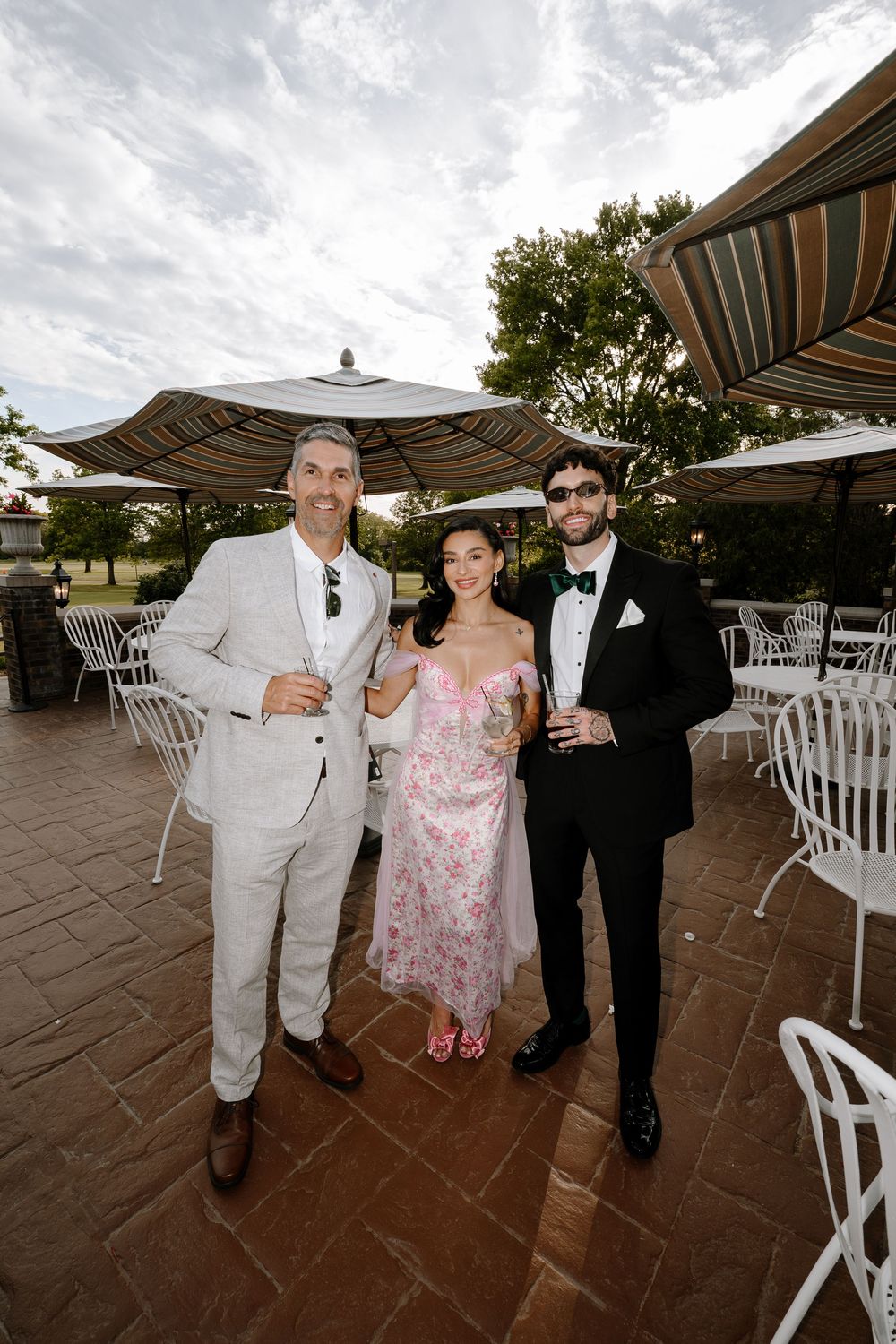 Two men and a woman in formal attire smile and toast drinks on a patio at a wedding reception.