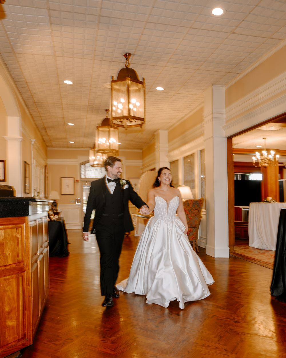 Bride in white ballgown and groom in black tuxedo walk hand-in-hand through elegant venue hallway with lantern lighting.