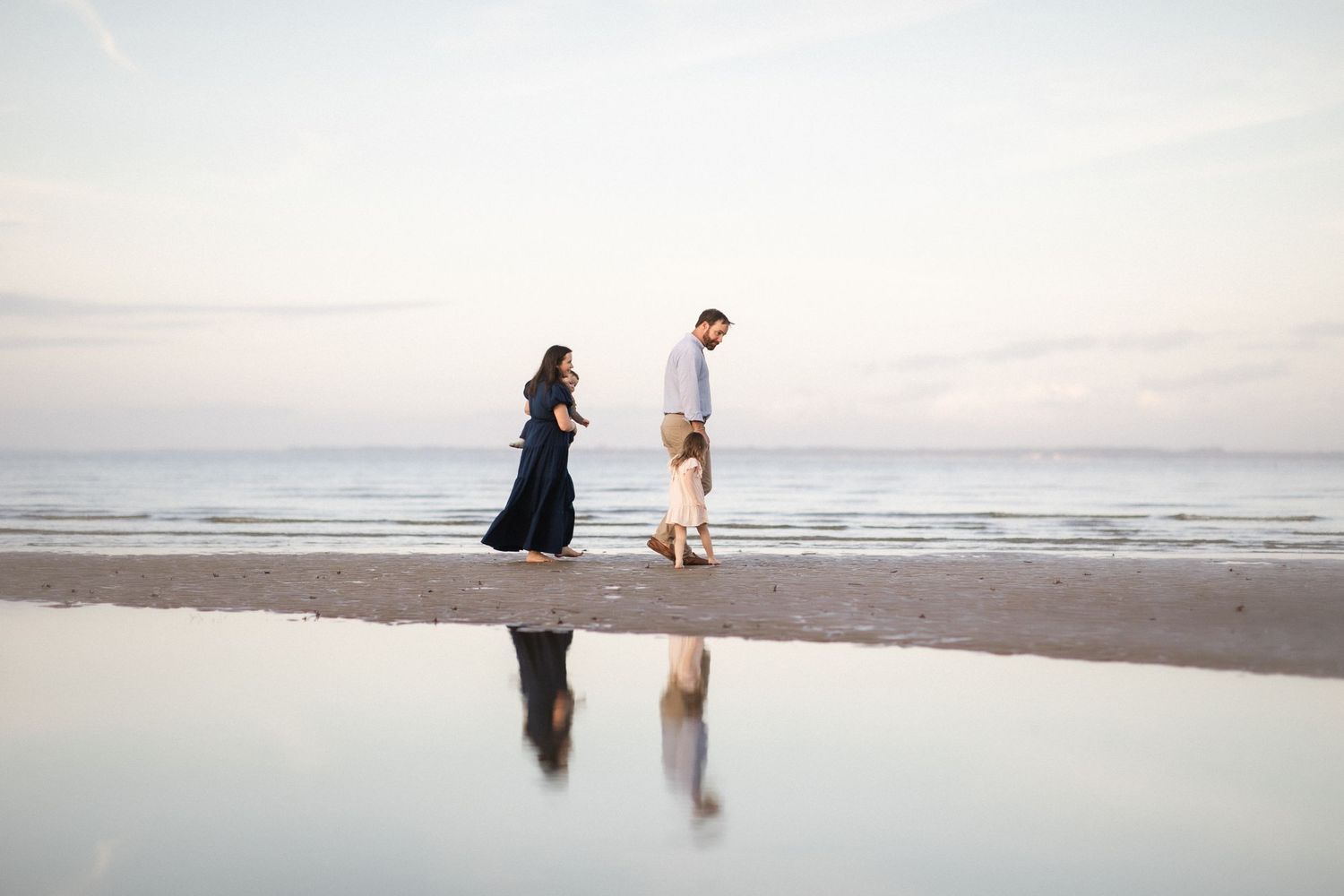Couple walking on a reflective beach at sunset, woman in navy dress and man in casual attire near ocean waves.