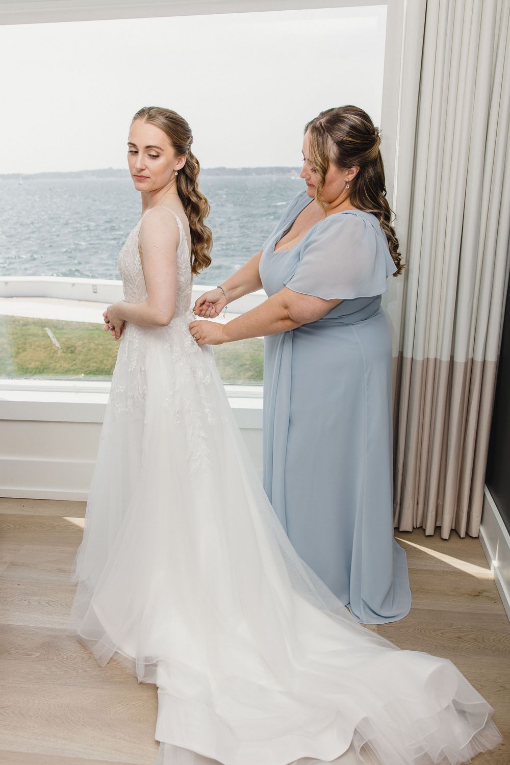 A bride in a white lace gown being helped by a woman in a blue dress near a window with ocean views.