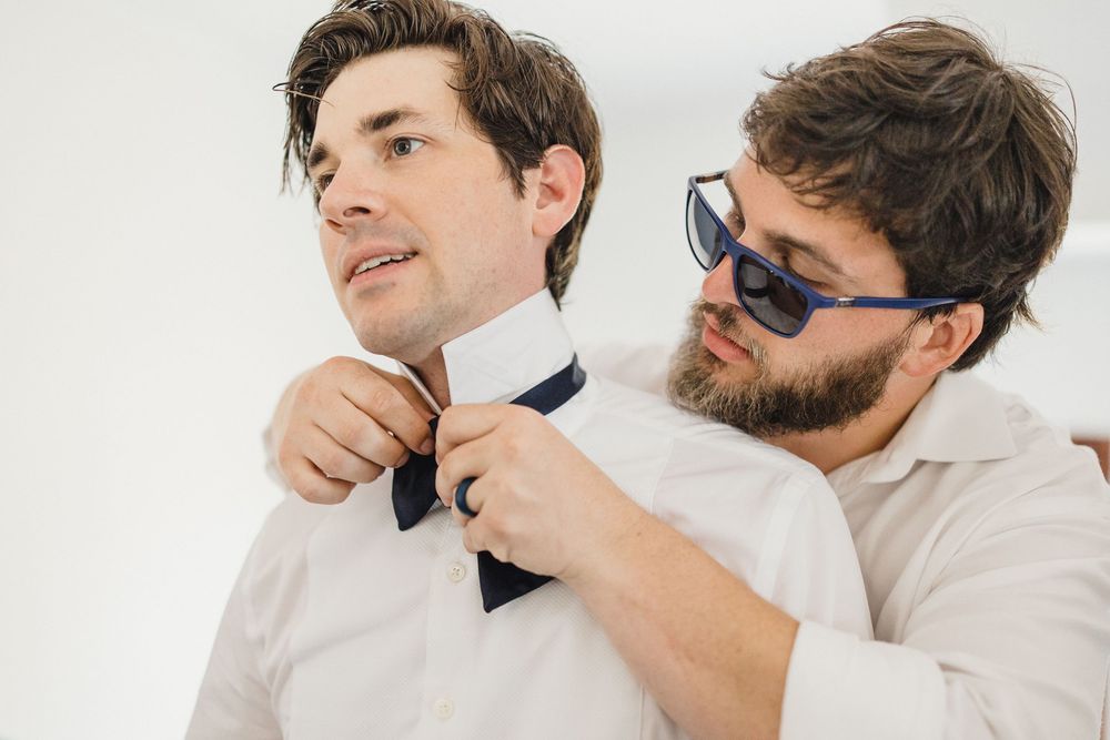 A bearded man in sunglasses helps adjust a navy bow tie on a well-dressed man in a white shirt.