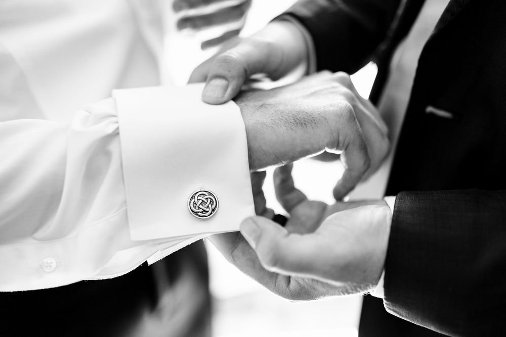 Black and white photo of a man fastening cufflinks on a white dress shirt sleeve at a formal event.
