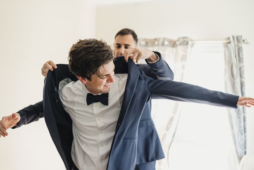 Groom being helped into his navy blue suit jacket by groomsman while wearing a black bow tie before wedding.