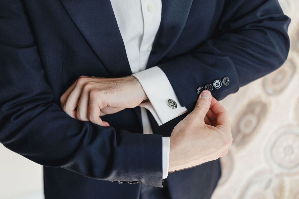 Man in navy suit adjusting silver cufflinks on white dress shirt, close-up of hands and wrists.