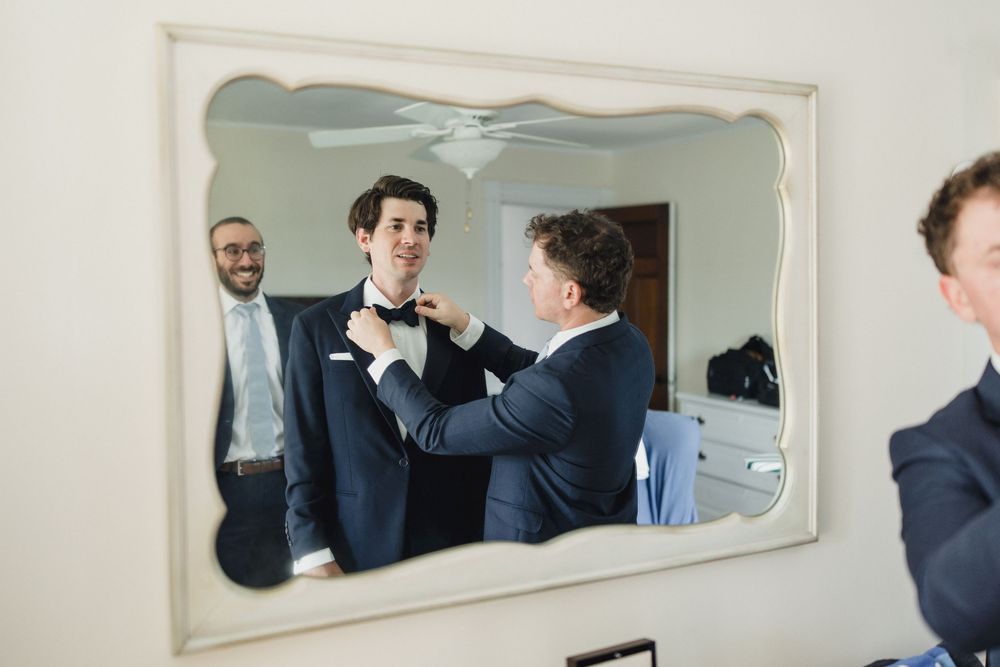 Groomsman adjusting bow tie of groom reflected in elegant white-framed mirror before wedding ceremony.