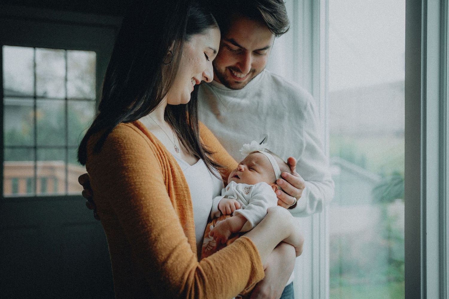 A young family sits on a bed, parents smiling as their baby explores a small object together.