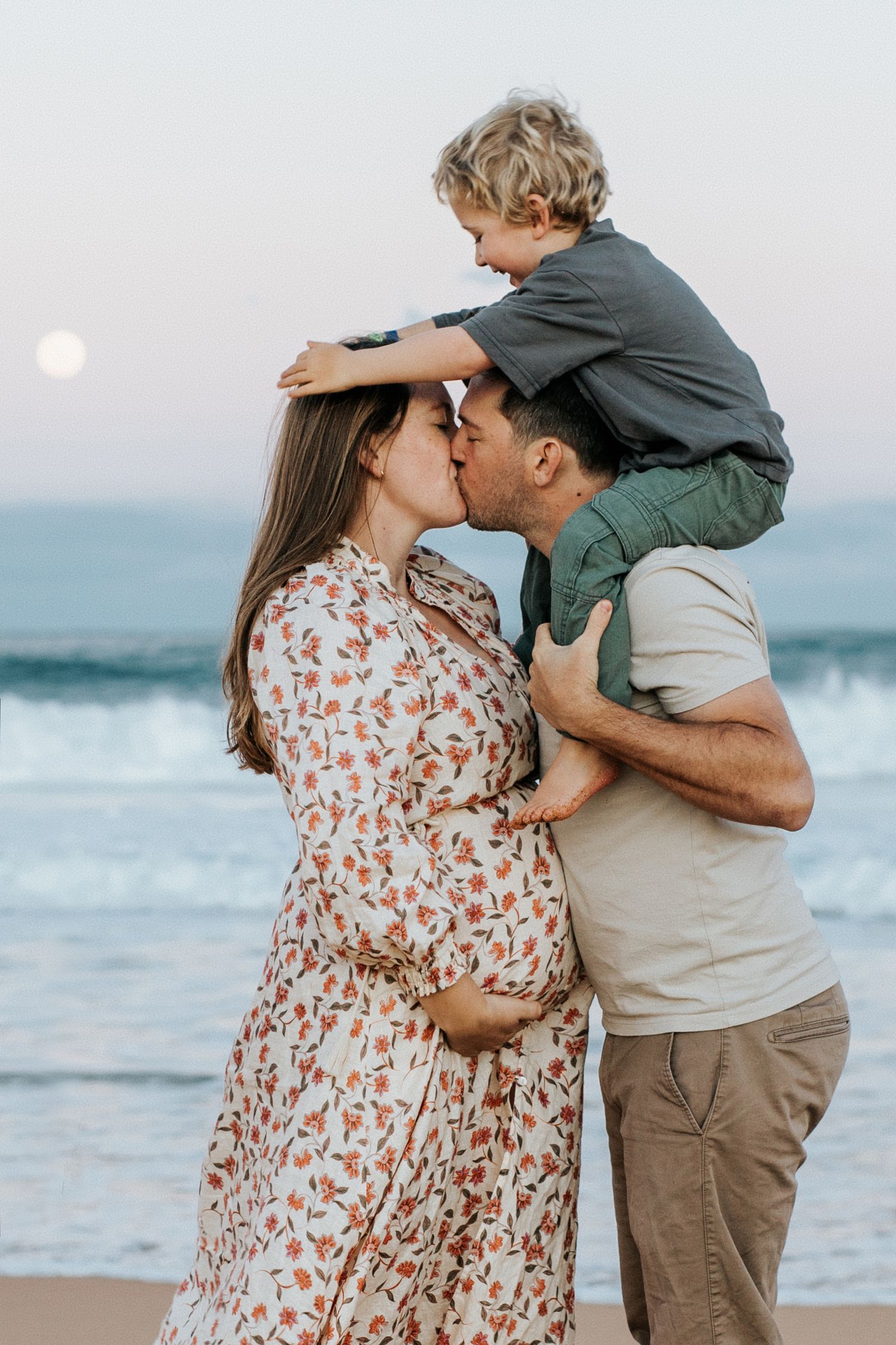 Pregnant woman in floral dress kissing partner on beach at sunset while young child sits on his shoulders.