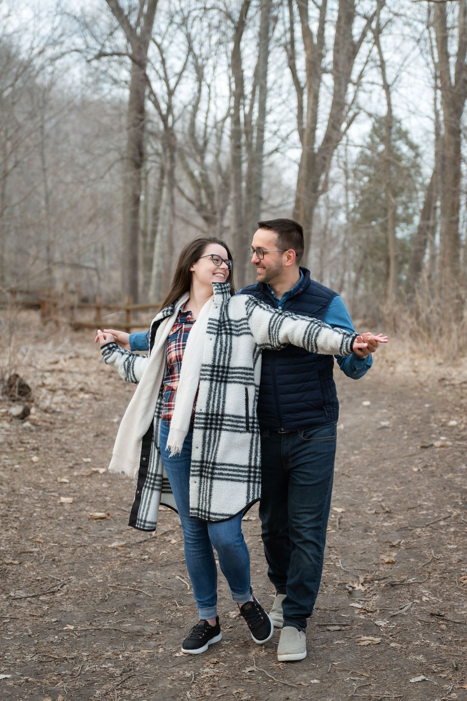 A smiling couple walks hand-in-hand on a wooded path in autumn, wearing plaid and puffer jackets.