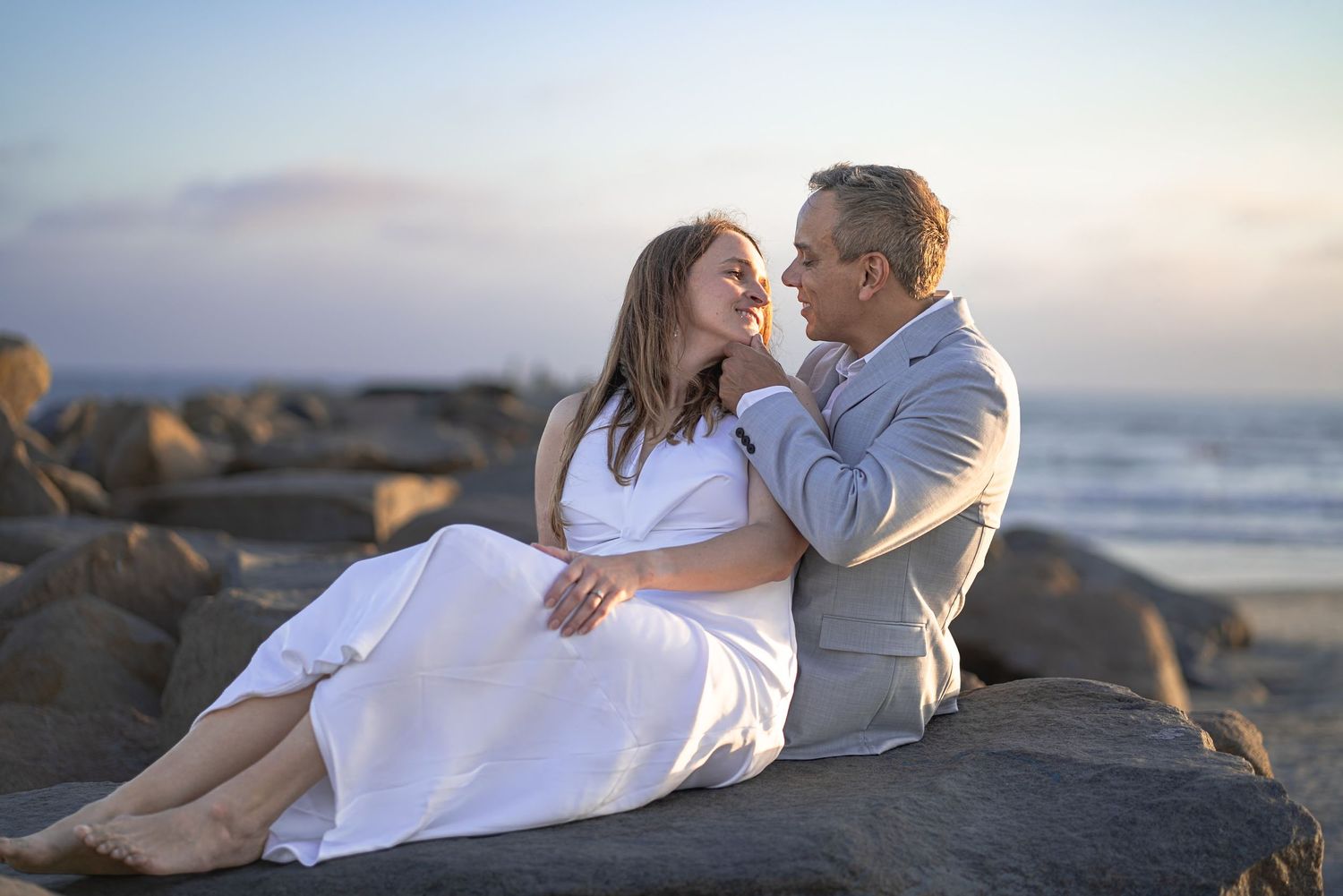 Photo of a newly married couple sitting on rocks by the ocean, gazing lovingly at each other. Oceanside, CA