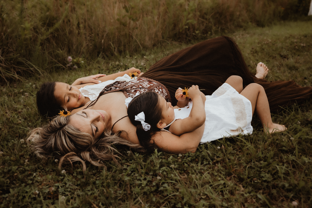 Mother and two young children lying together in green grass, sharing a tender moment outdoors.