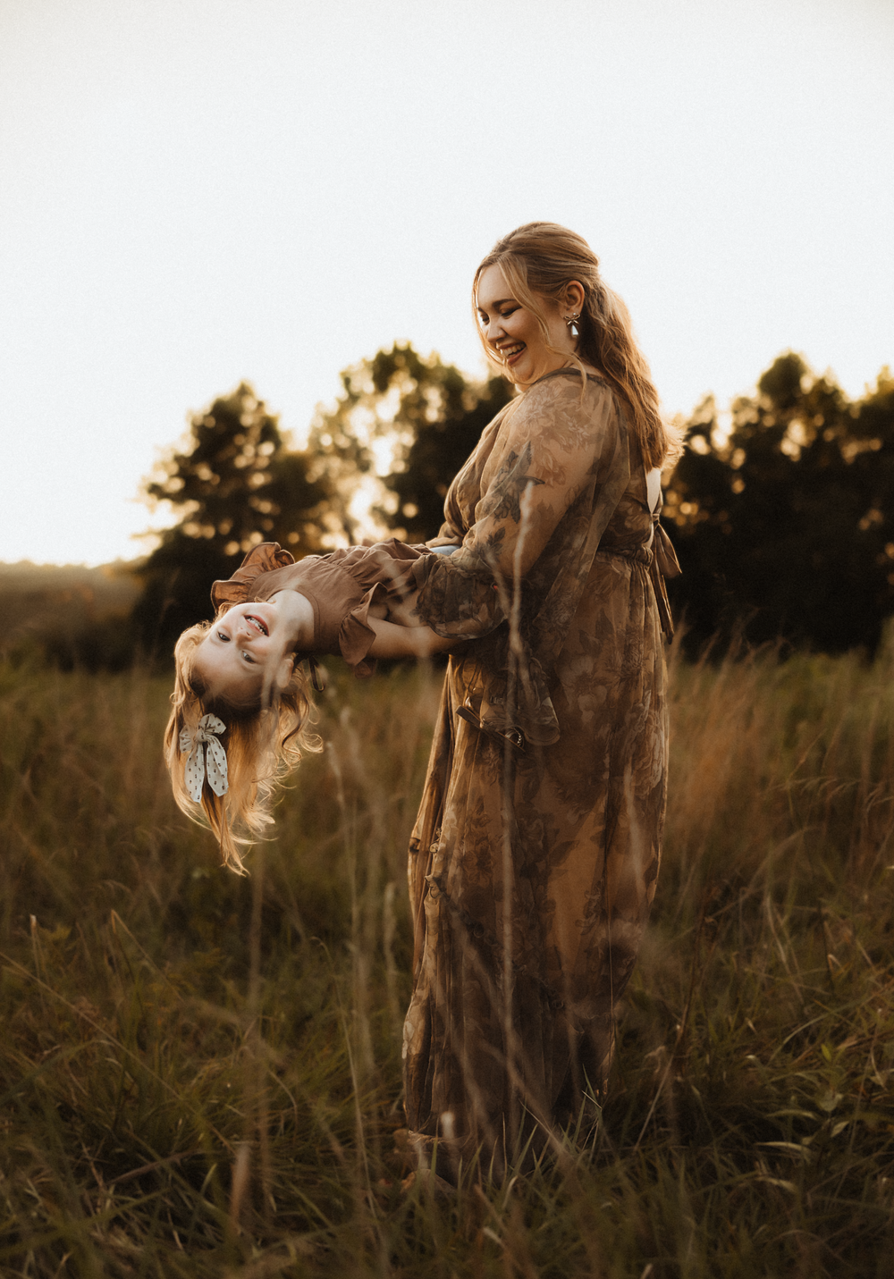 A smiling woman in a boho floral dress lifts a laughing child upside down in a golden sunlit field.