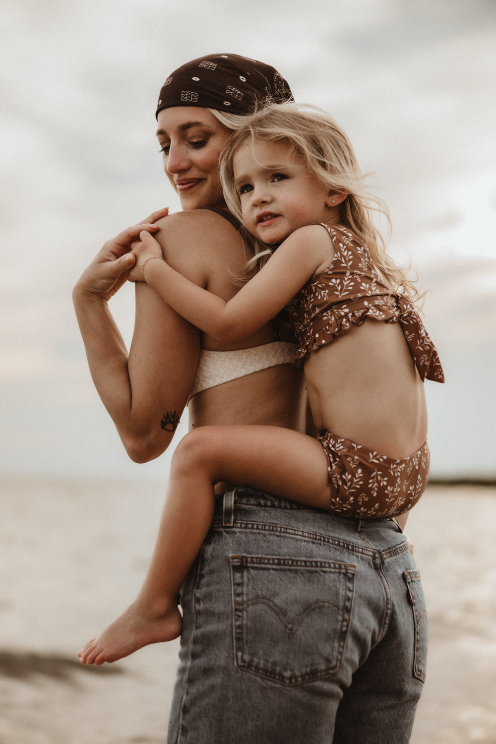 A mother holds her young blonde daughter on a beach, both wearing brown swimwear, sharing a tender embrace.