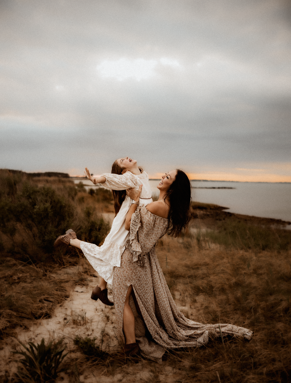 Two women in boho dresses joyfully dancing on a sandy coastal dune at sunset.