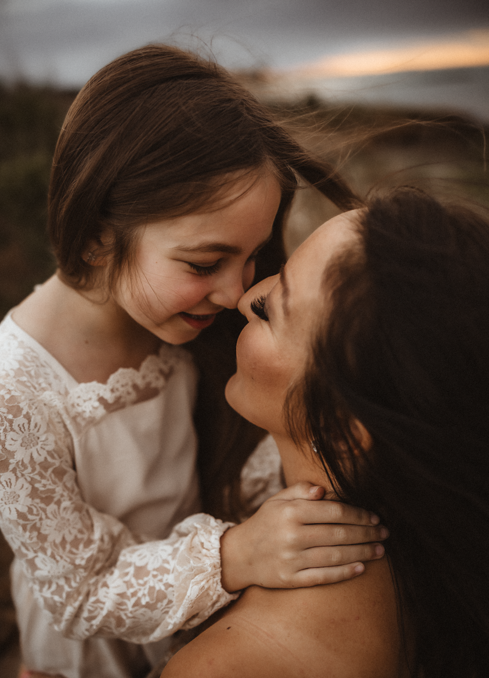 A mother and young daughter share a tender nose-to-nose moment, smiling warmly during a golden hour outdoor portrait.