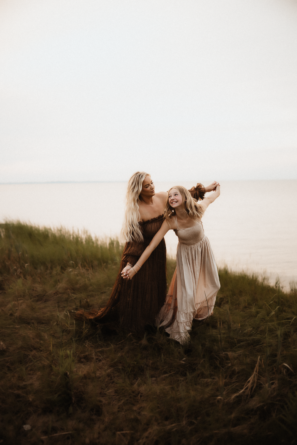 Two women in flowing boho dresses dance joyfully in a grassy field near water at golden hour.