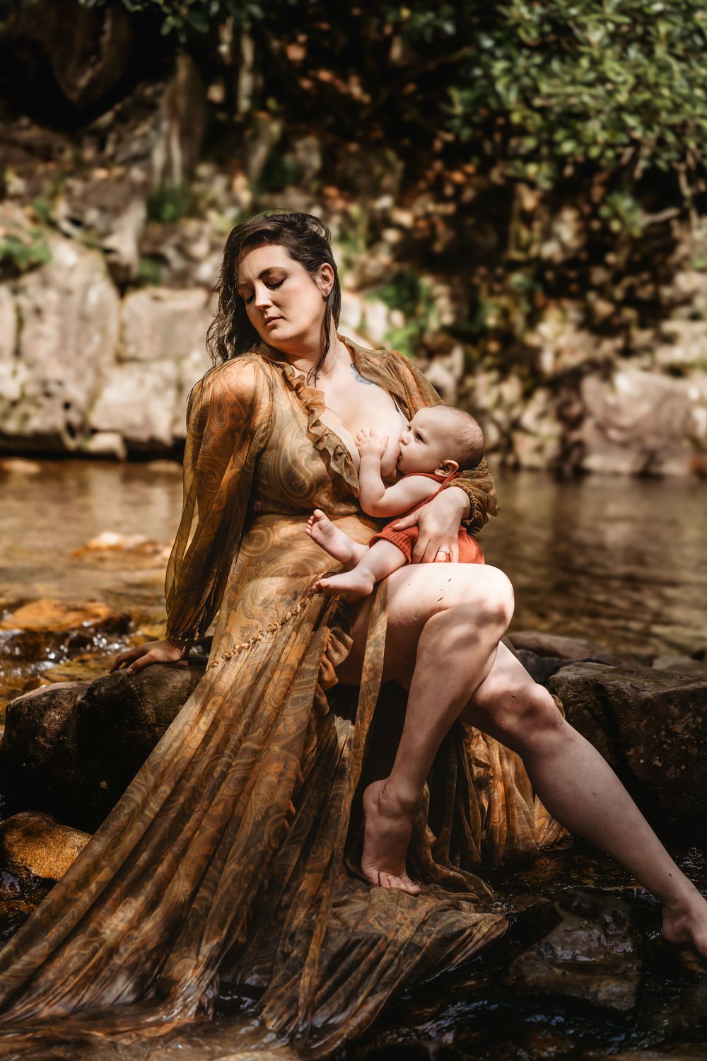 Artistic motherhood portrait by river with woman in gold dress holding baby.