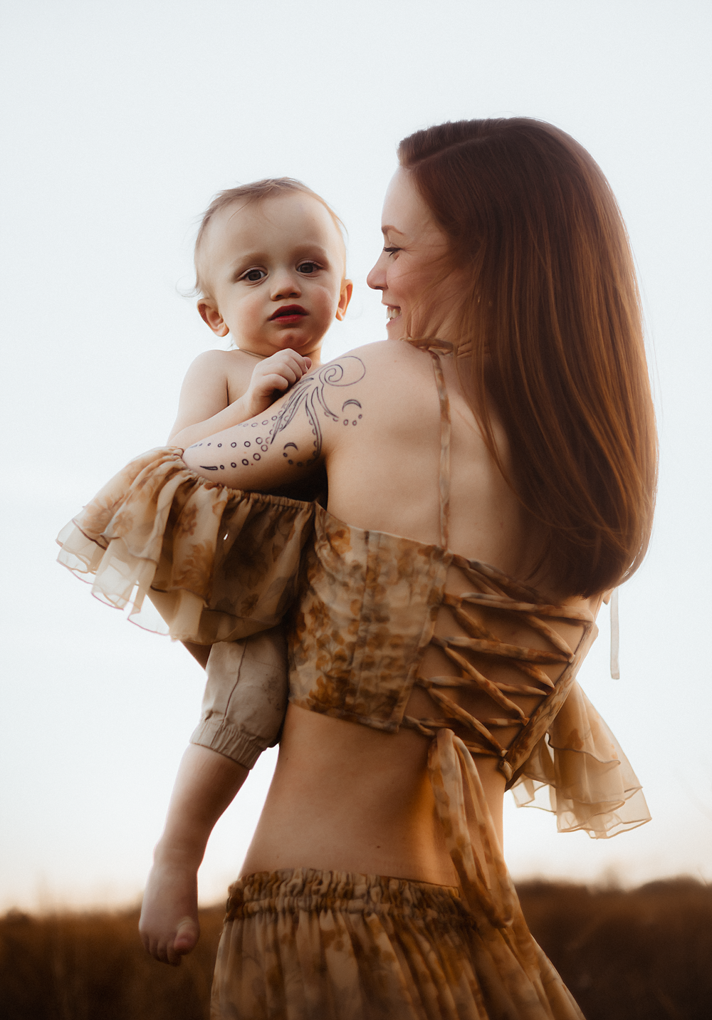 A tattooed mother in a brown off-shoulder dress holds her smiling baby outdoors in warm golden light.