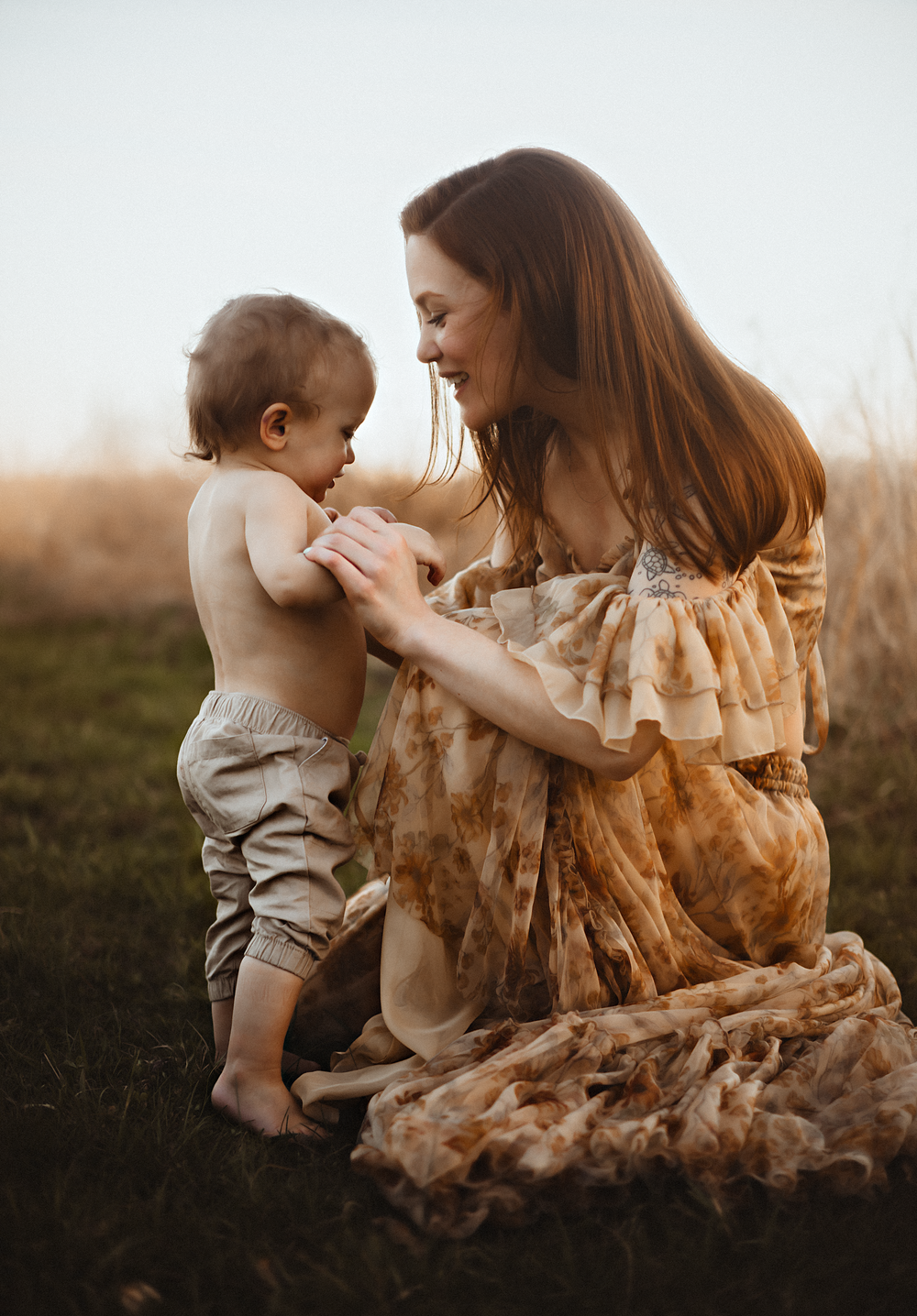 A smiling mother in a floral dress sits on grass, tenderly holding her toddler in golden hour light.
