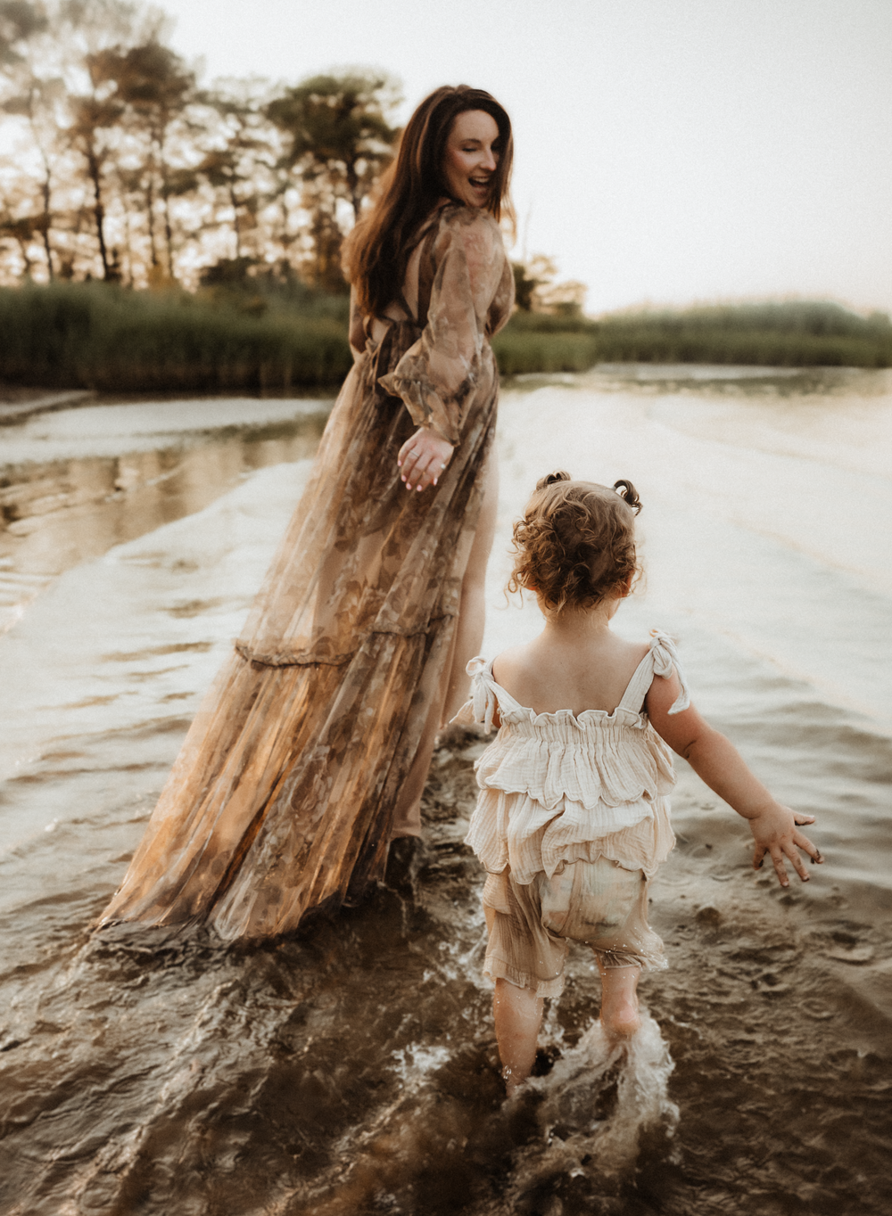 A mother in a floral maxi dress holds her toddler's hand while wading through shallow water at golden hour.