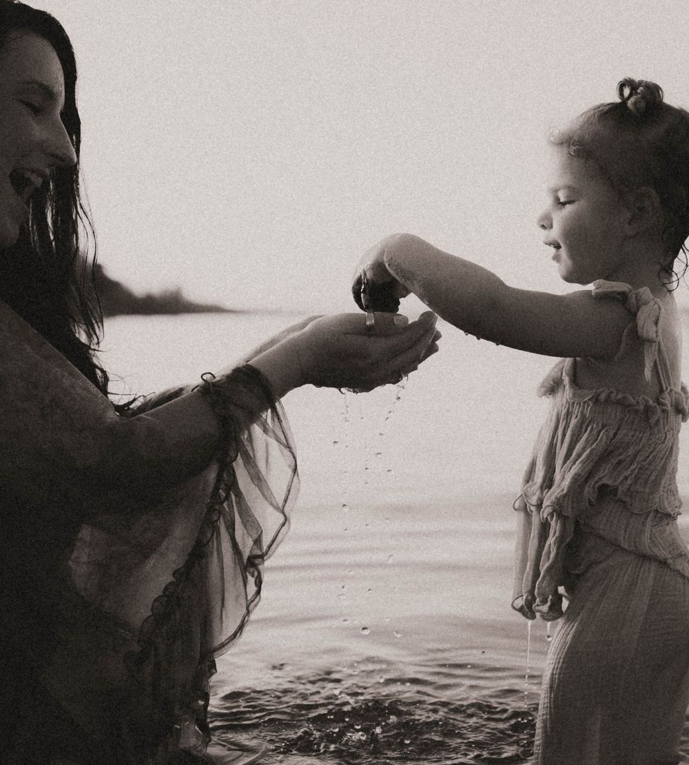 Black and white photo of a mother and young daughter playing with water by a lake shore.