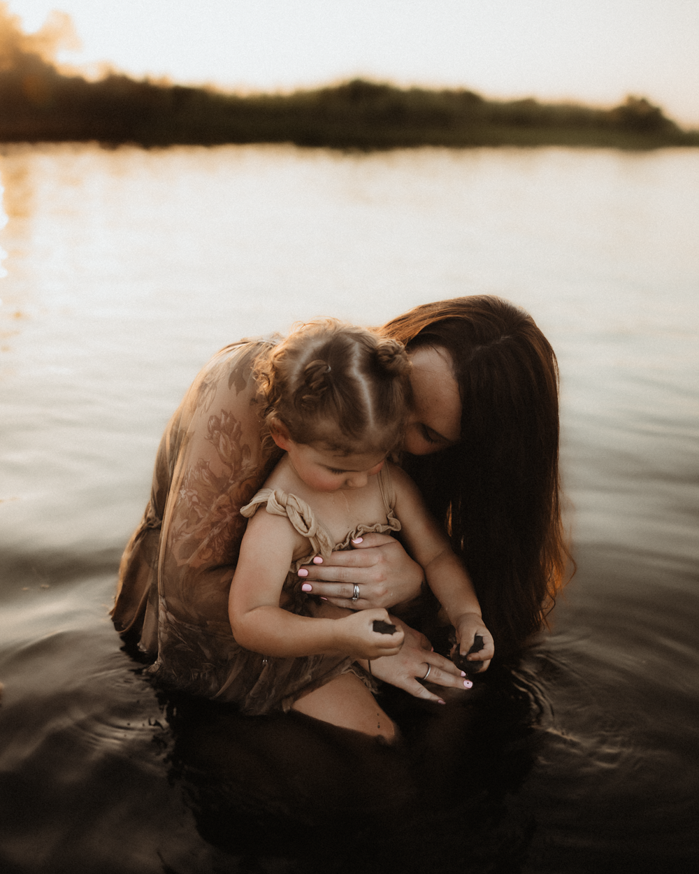 A mother tenderly holds her toddler while standing in a calm lake at golden hour sunset.