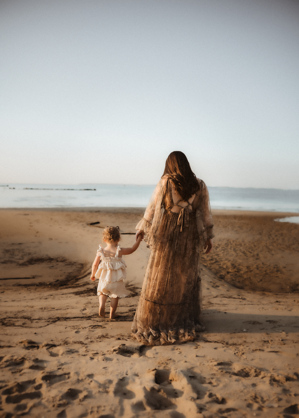 A mother and young daughter walk hand-in-hand on a sandy beach toward the ocean, woman wearing a flowing boho gown.