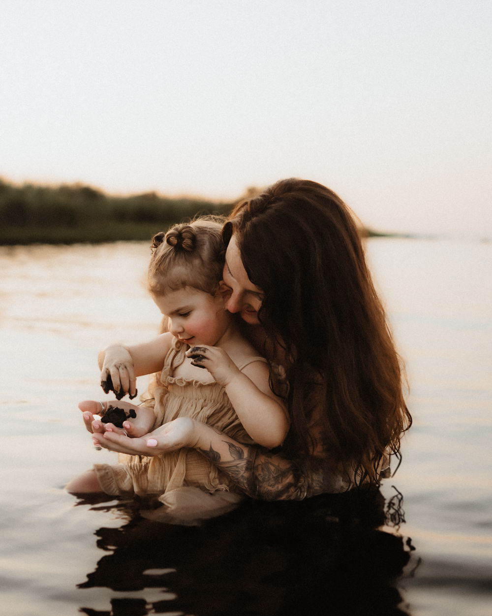 Mother and young daughter sitting in calm water at sunset, sharing a tender moment together.