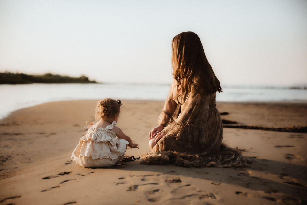 Mother and young daughter sitting together on a sandy beach at sunset, viewed from behind.